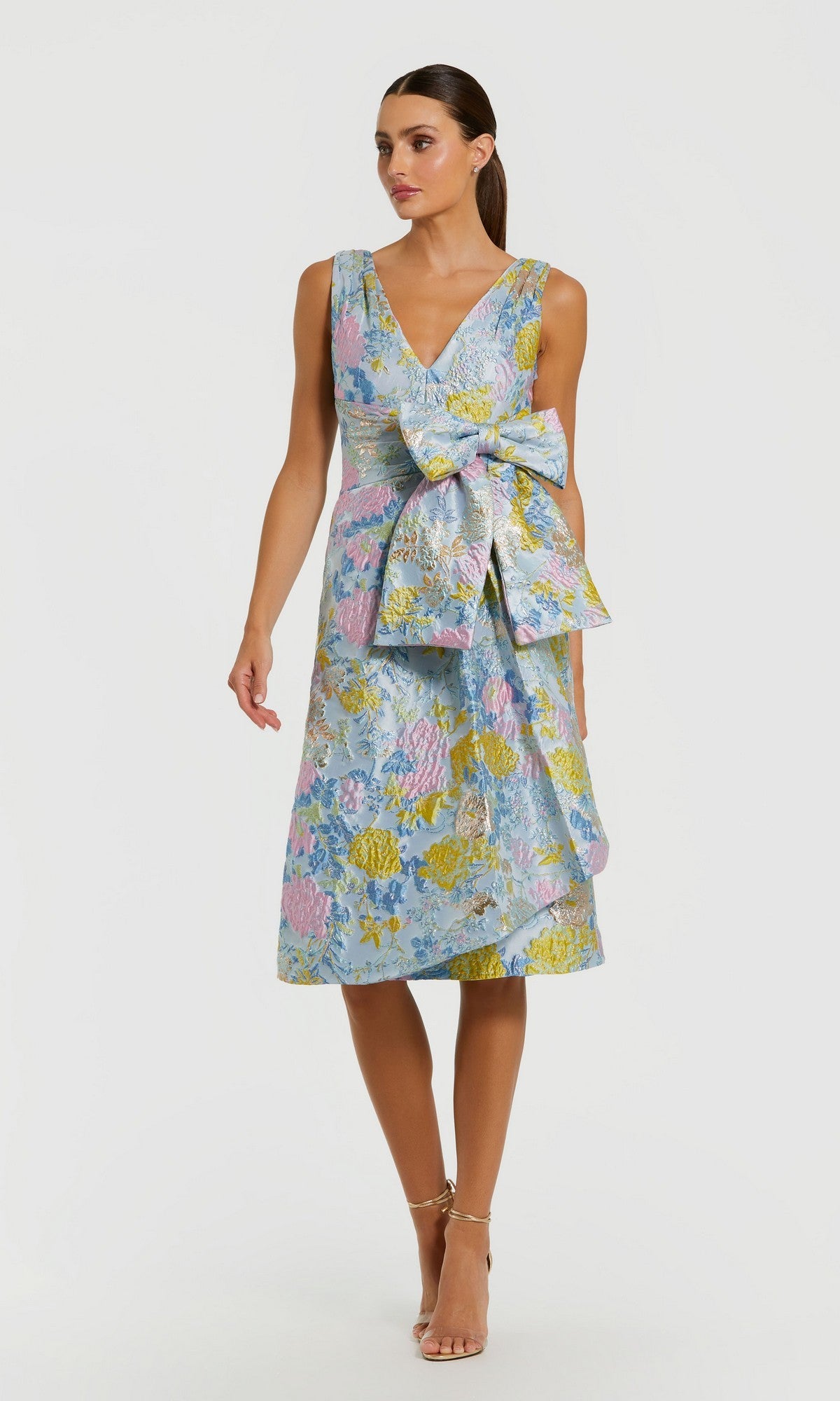 Woman wearing the Mac Duggal 56104 knee length wedding guest dress in Blue Multi, featuring a sleeveless floral brocade design with a large waist bow, standing against a plain white background.
